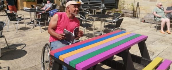 A wheelchair user sitting at a multicoloured Bradshaw recycled plastic picnic table on the cafe terrace at Hebden Bridge Town Hall
