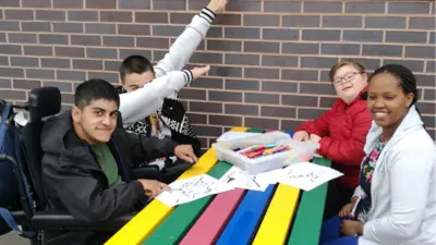 Young people drawing, smiling, laughing at colourful table. Includes wheelchair users. British Recycled Plastic