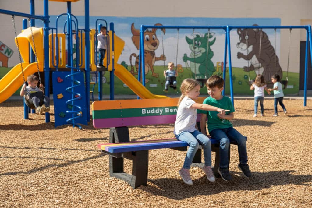A smooth-corner Buddy Bench made from durable, 100% recycled British plastic, designed for safety, comfort, and long-lasting use in schools and play areas. 2 children sat on bench smiling and pointing