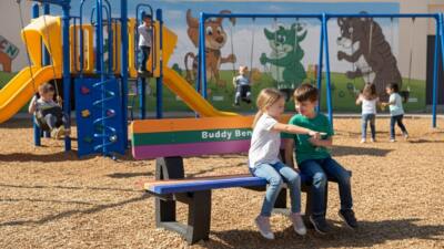 A smooth-corner Buddy Bench made from durable, 100% recycled British plastic, designed for safety, comfort, and long-lasting use in schools and play areas. 2 children sat on bench smiling and pointing