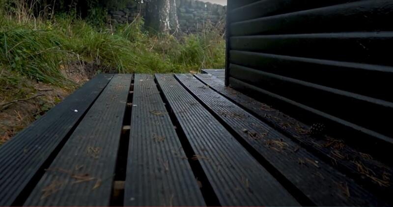 Artificial lumber decking outside a black wooden structure, surrounded by grass and a stone wall, with scattered pine needles