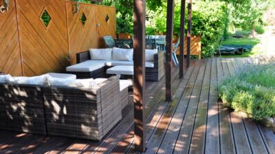 Outdoor patio with artificial lumber decking, rattan seating with white cushions, a wooden pergola, and lush greenery in the background