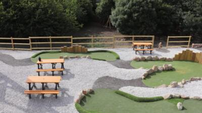 Outdoor mini-golf course with artificial grass and gravel paths, fencing, and recycled plastic picnic tables with orange tops and seats.