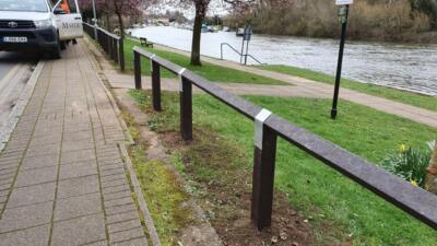 Recycled plastic bird's mouth fencing installed along a riverside path, with a river, trees, and a footpath visible in the background.