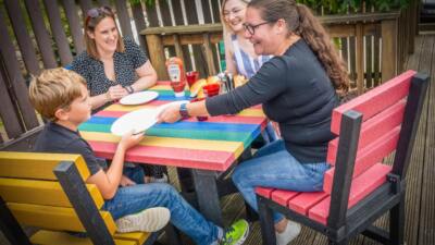 A family of four dining outdoors at a colourful recycled plastic table with rainbow-coloured chairs.