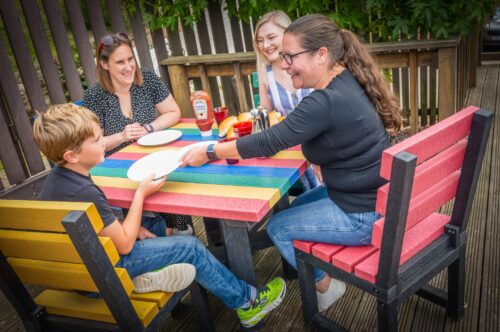 A family of four dining outdoors at a colourful recycled plastic table with rainbow-coloured chairs.