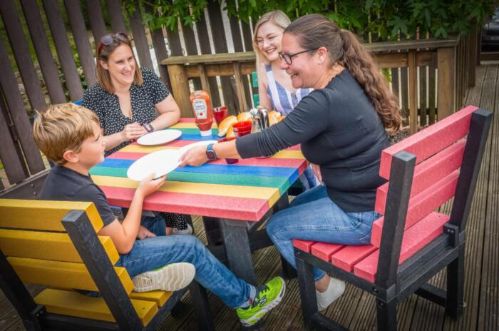 FEatherstone Square in Colour with three adults and child A family of four dining outdoors at a colourful recycled plastic table with rainbow-coloured chairs.