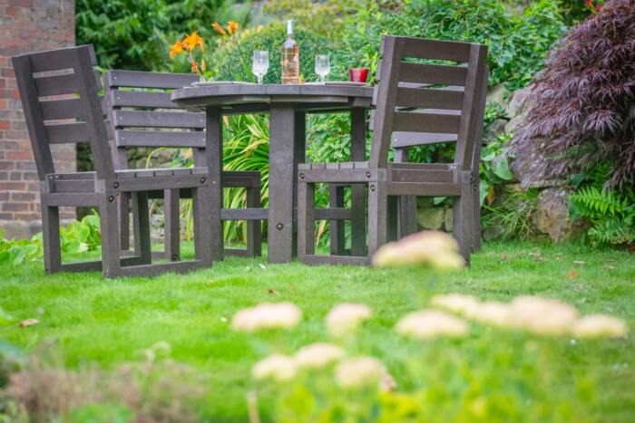 Featherstone Round in Brown from below with flowers Recycled plastic dining set in a vibrant garden with a round table, four chairs, and a bottle of wine for an inviting outdoor setting