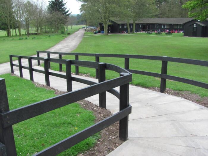 Post and rail 1 Pathway with black recycled plastic handrails winding through green grass towards a building in a park setting