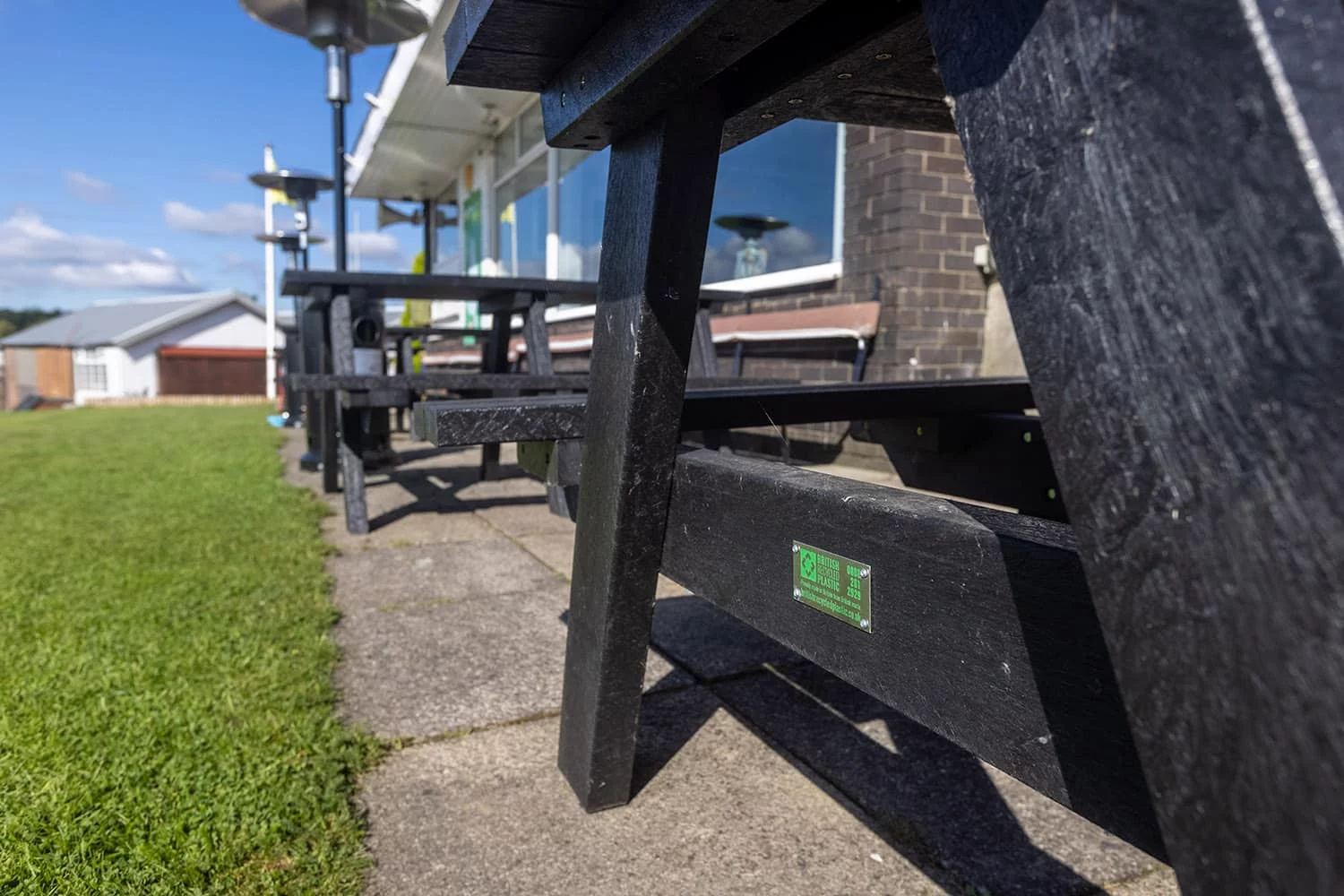 close up of black artificial lumber that has created a British Recycled plastic bench that are situated at a sports club. You can see the quality logo mark on the bench