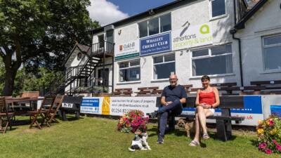 2 people sat at a black British recycled plastic bench. its in a cricket club garden as there is a sign behind that says 'whalley cricket club'