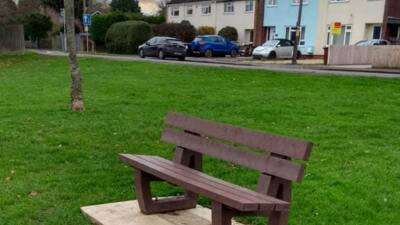 A brown british recycled plastic bench in a small patch of grass with houses nearby