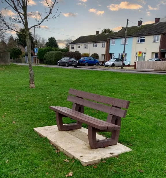 A brown british recycled plastic bench in a small patch of grass with houses nearby