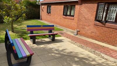 2 rainbow coloured British recycled plastic benches adjacent to each other next to a brick house.