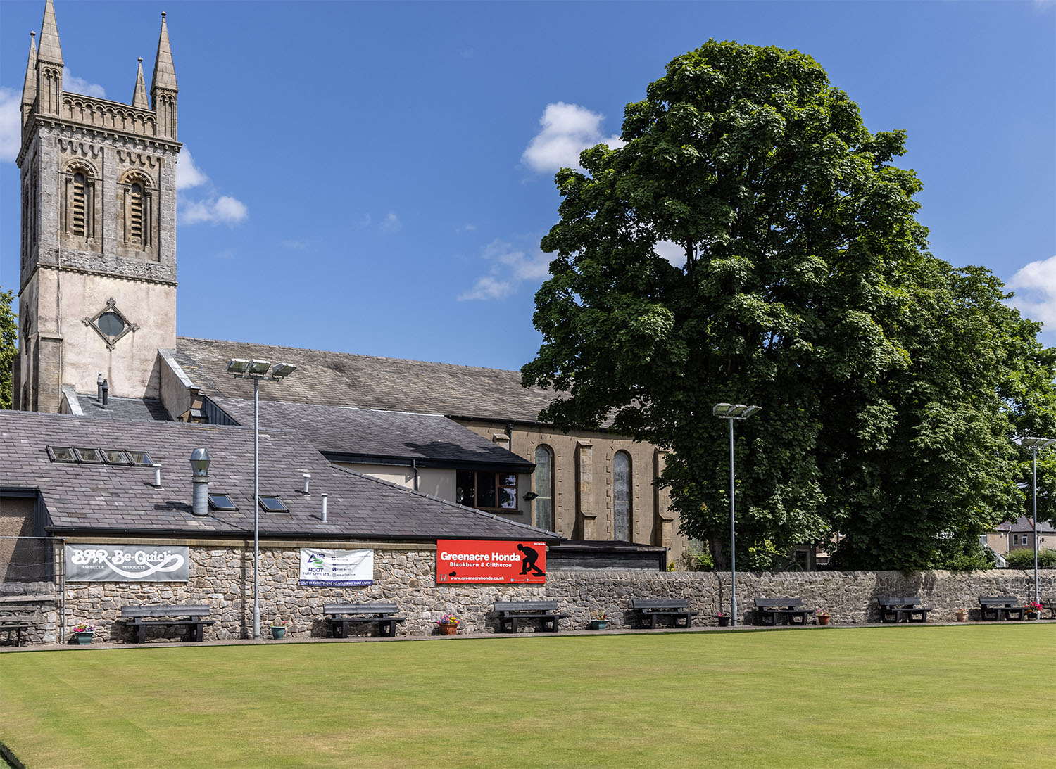 The Plastic Problem We Don't Talk About: Artificial Grass & Microplastics 3 a shot of 8 black British recycled plastic benches next to a beautiful church and a bowling green in a beautiful english village