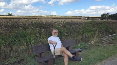 a very smiley man sat on a british recycled plastic bench holding a spade