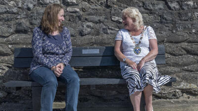 2 ladies sat on a black British recycled plastic bench. They are smiling at each other