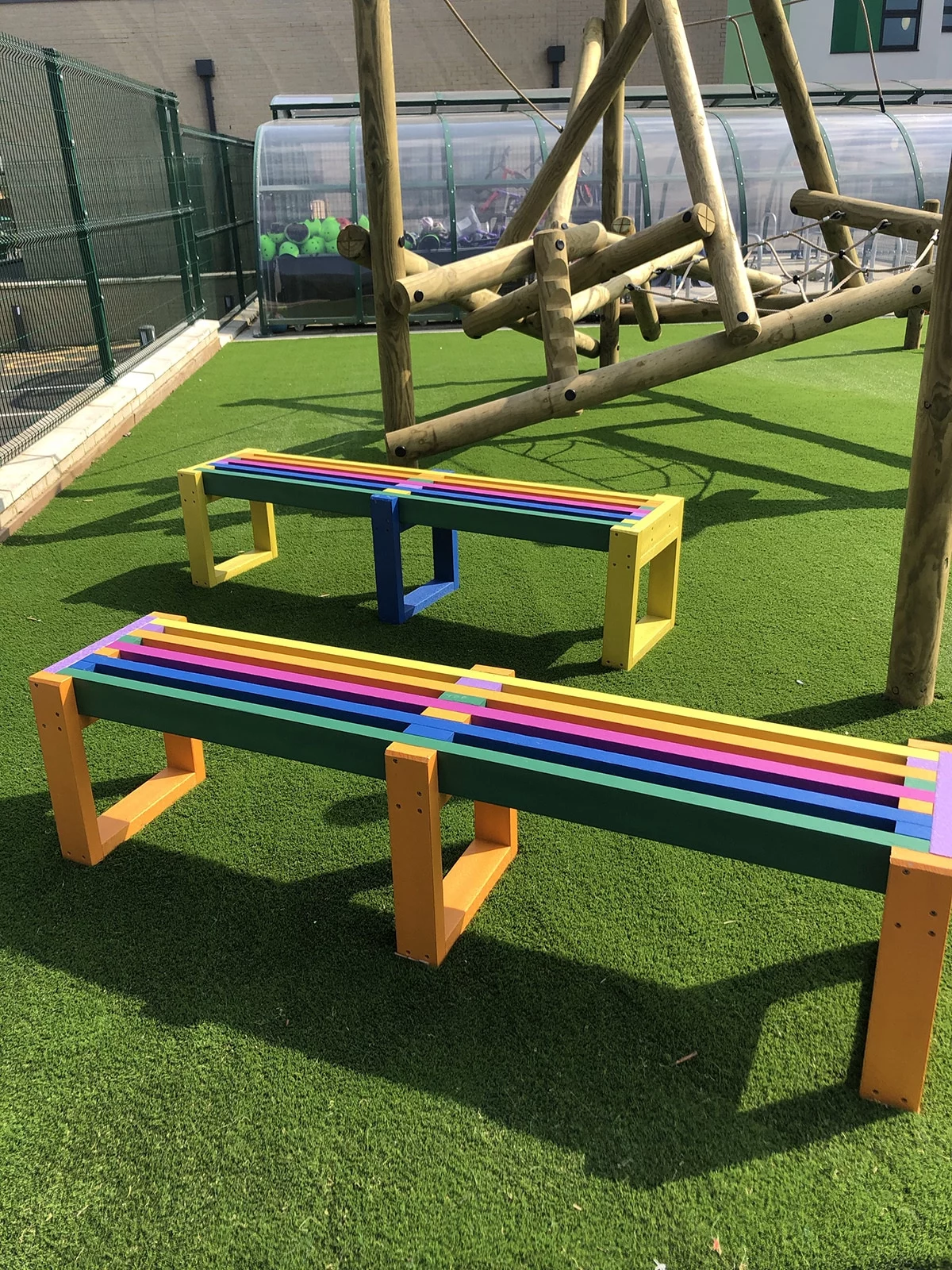 2 backless British recycled plastic benches that are rainbow coloured. They are in a pub beer garden and there is a wooden playground nearby