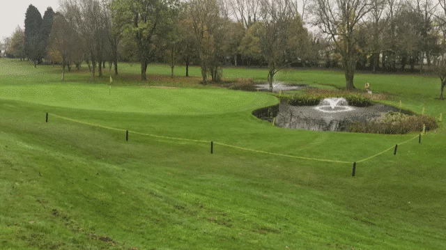 Golf course with lake and water fountains, surrounded buy bright blue sky and lush green trees.