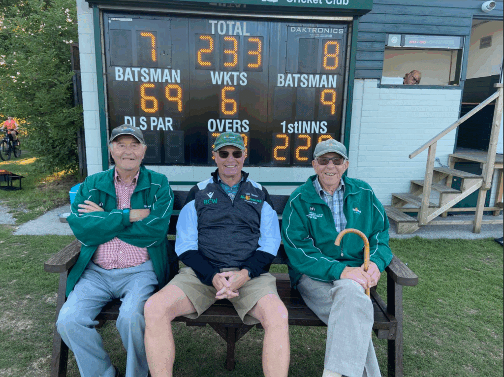 Cricket club with 3 men smiling, sat on our Harewood bench with the cricket score board in the background