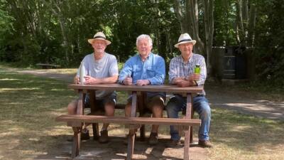 3 men sat smiling on a brown picnic table with woodland in the background