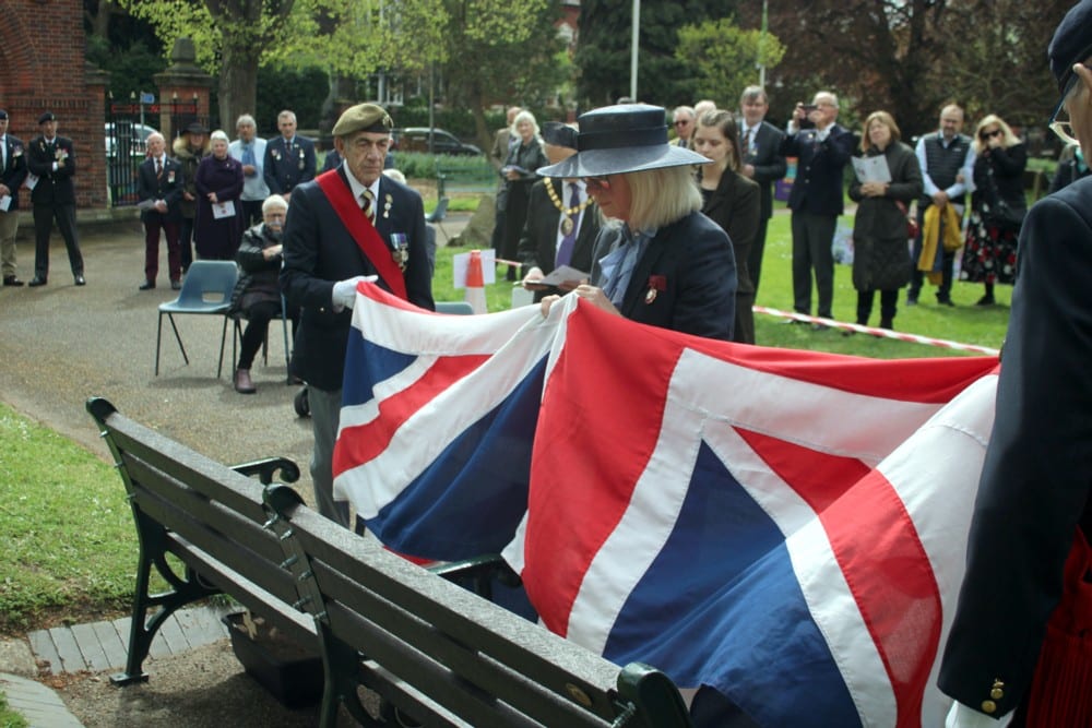 Community Memorial Bench Restoration in the Heart of Worcester 4 Veterans holding a Union Jack flag unveil a memorial bench in Gheluvelt Park, Worcester, with members of the public stood watching.