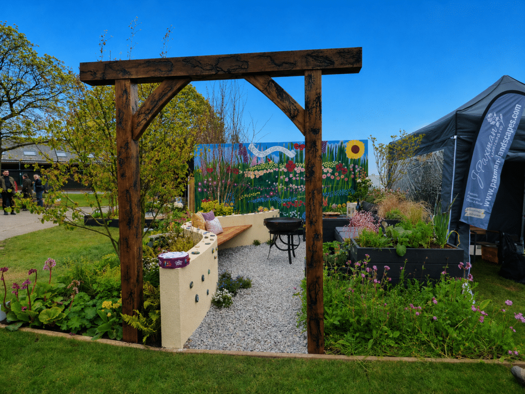A front-facing view of Common Ground, a community show garden created for the Harrogate Flower Show. A large wooden archway is surrounded by a variety of flowers and plants. Behind them is a low curved wall embedded with hand-painted pebbles, plastic planters, and a large painted mural which says 'COMMON GROUND 2026'.