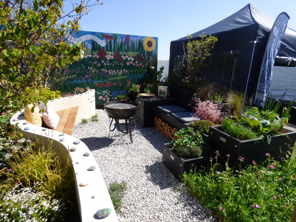 An interior view of the Common Ground show garden. A low curved wall is embedded with hand-painted pebbles. Directly across are plastic planters and seating, which are surrounded by plants and vegetation. A fire pit sits in the middle of the garden on top of the gravel flooring.