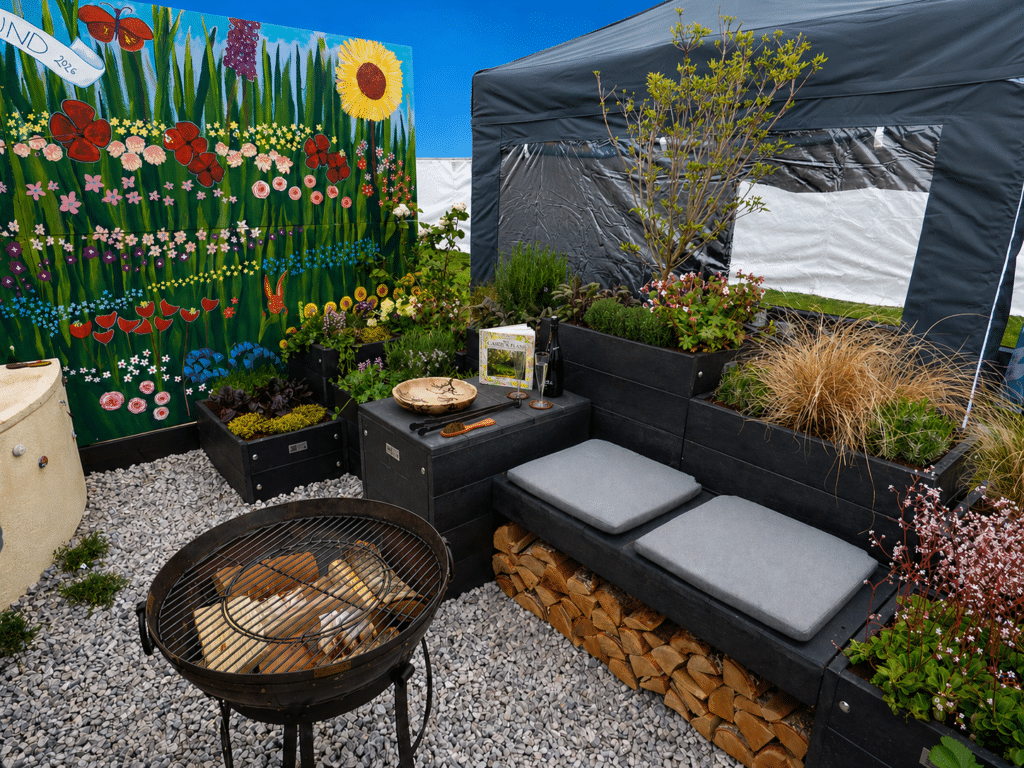 An interior view of the show garden Common Ground. A fire pit stands in front of recycled plastic seating and planters which are filled with a variety of plants. The display is backed by a large, hand-painted mural of flowers in different colours.