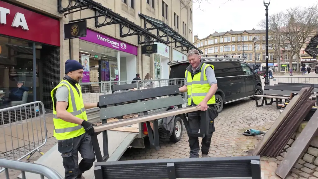 Two men, both wearing grey t-shirts, black pants, and yellow hi-vis vests, carry one of the recycled plastic benches ready to be set up outside the Woolshops Shopping Centre in Halifax, West Yorkshire, UK.