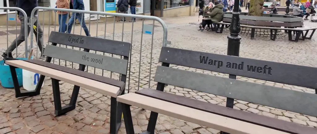 Two sections of the Calverley outdoor benches, made with recycled plastic slats in grey, brown, black, and beige. The tree benches have phrases etched into them, themed around Halifax, Yorkshire and its history.